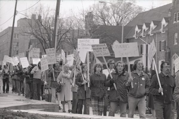 View towards large group of people marching with signs on the sidewalk at the office of State Superintendent of Education, Barbara Thompson at the Department of Public Instruction. The protest is in support of striking Hortonville teachers. Madison Mayor, Paul Soglin, can be seen at the back of the group at left.