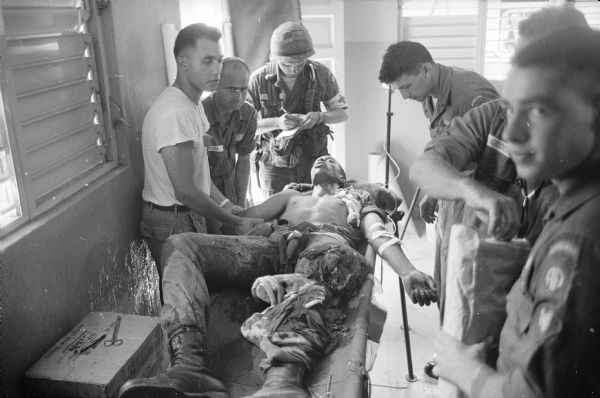 Medics treating Paratrooper Private First Class Richard F. Greene of Silver Springs, MD at Airborne battalion aid station at Santo Domingo, Dominican Republic. Greene's leg was shot off at the thigh. Name tags of medical personnel that are visible include Giddens and Dixon.