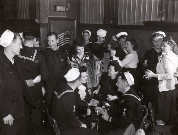 Two sailors and two women are seated at a table smoking cigarettes and drinking beer, while another woman stands playing an accordion for them. Around the table are other sailors and women, with a number of couples dancing.