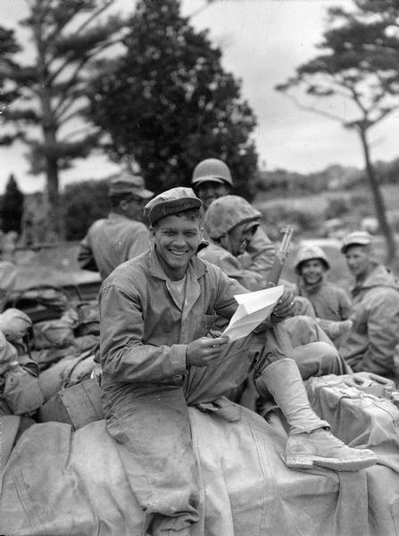 Soldier L. Heims, seated outdoors on the back of a loaded vehicle, smiling broadly while reading a letter. Other smiling soldiers are in the background.