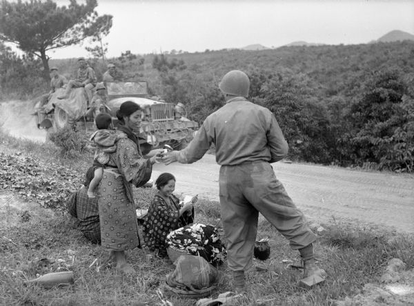 Soldier offering food to Japanese women and children gathered on a roadside in Okinawa. One woman wears a baby on her back. Other soldiers watch from a jeep on the road in the background.