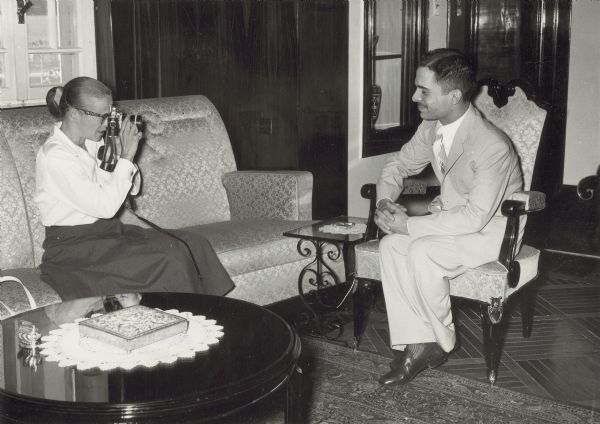 Dickey Chapelle seated on a sofa photographing King Hussein, who is seated across from her in a chair.