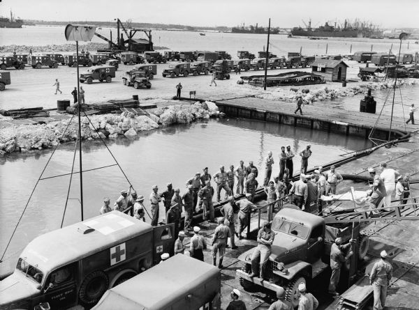 Elevated view looking down towards a group of men taking a wounded soldier down a ramp from the USS <i>Samaritan</i> to a waiting ambulance. Several military personnel stand by on the dock. A fleet of ambulances are parked in rows across a river or canal on the other side of the dock. More ships are in the far background across the water.