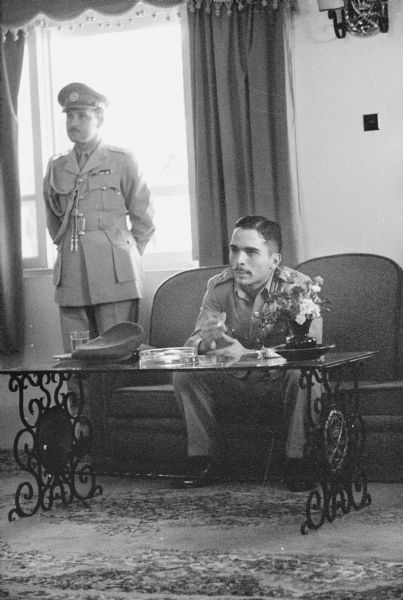 King Hussein of Jordan seated on a couch behind a small table. A man in military uniform stands beside the king on the left.