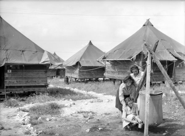 Three women getting water at a reservoir in the 148th General Hospital compound at Saipan. Several raised tents can be seen in the background.