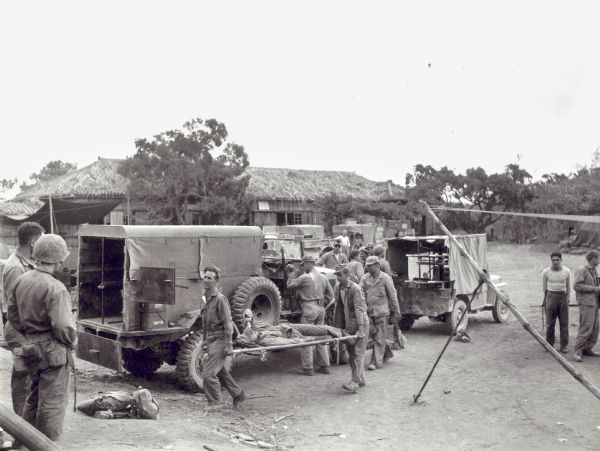 Two men carry a wounded soldier on a stretcher toward the back of a waiting vehicle to transport him to another location. A grass-roofed building is in the background.