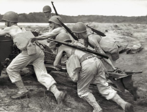 Four shirtless soldiers strain to push a large gun over sandy terrain in Panama. Each man also wears a helmet and carries a backpack, a rifle, and a canteen. Another soldier walks alongside the working group.