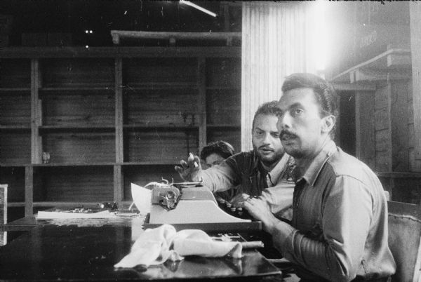 Three Cuban rebels seated at a table. One of the men is using a typewriter.