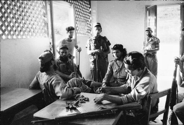 Group of soldiers backing Fidel Castro in the Cuban Revolution gathered in a small room with open doorways. One man is seated at a table writing, while other men sit and stand around the room holding weapons. One of the seated men, in the center, is probably Camilo Cinfuegos.
