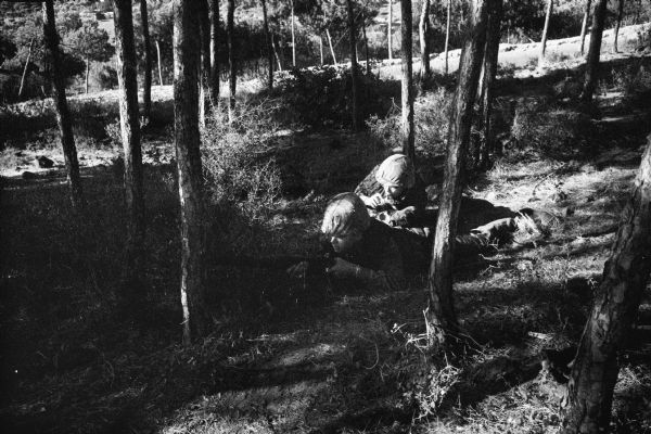 Dickey Chapelle in Lebanon wearing camouflaged clothing and a helmet crouched low to the ground and holding a camera. She is next to and behind a soldier who is laying flat on his stomach in a sparsely wooded area with a rifle aimed to the left.