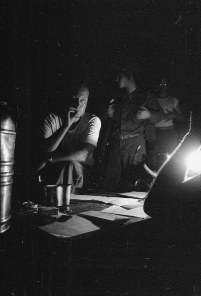 View at night in Lebanon of a man sitting behind a table and smoking. He is illuminated by a lamp on the right. Other men stand behind him. In the foreground on the right is the arm of a man sitting at the table which is covered with papers.