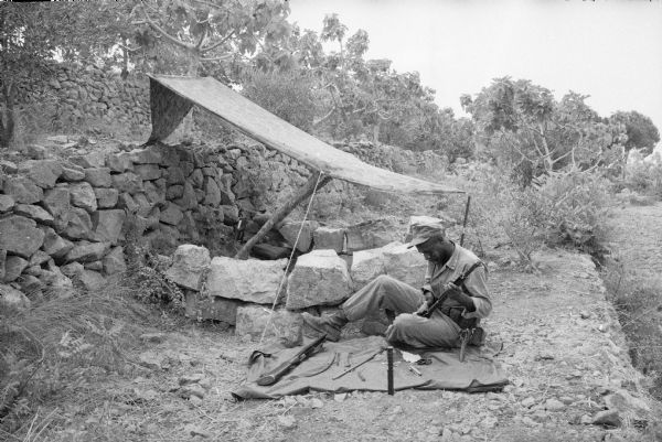 Lebanese soldier cleaning his rifle. He is seated on a blanket on the ground in front of a lean-to attached to a stone wall over a small stone bunker.