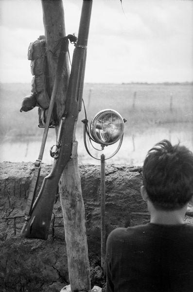 Rear view of a soldier at a wall on the edge of the Vietnamese village of Binh Hung. A rifle and pack are hanging from a wooden post, and next to it is a spotlight mounted on a metal post.