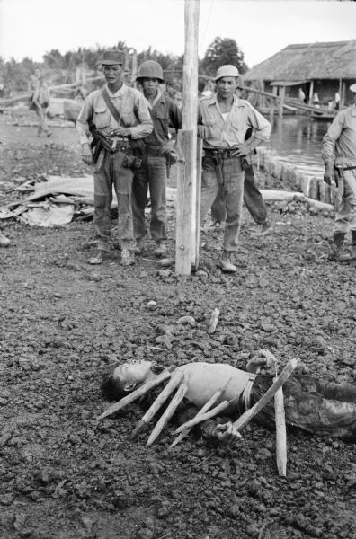 Dead body of a Viet Cong soldier on display in the marketplace of the Tan Hung village. He was caught planting poisoned spikes in swamps. Vietnamese soldiers are looking on in the background.