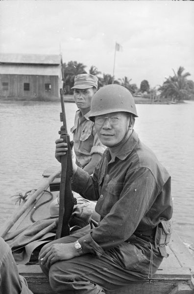 Father Augustin Nguyen lac Hoa, the fighting priest, sits in a boat on the canal between Binh Hung and Tan Hung Tay. Father Hoa is in uniform and holds a rifle while another soldier pilots the boat. Another man sits in the boat behind him. A building and a flag are on the far shoreline in the background.