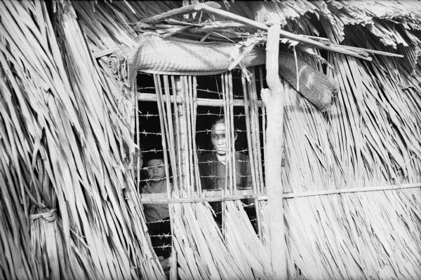 Two Viet Cong prisoners seen through a window covered in barbed wire in a structure built of bamboo and grass.