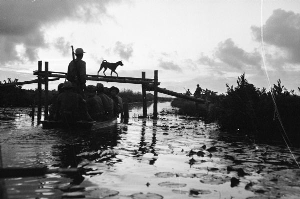 Silhouetted view across water of a group of people on a small boat near the bridge on the canal at Tan Hung Tay, Vietnam. A dog is crossing the bridge and soldiers patrol in the background.