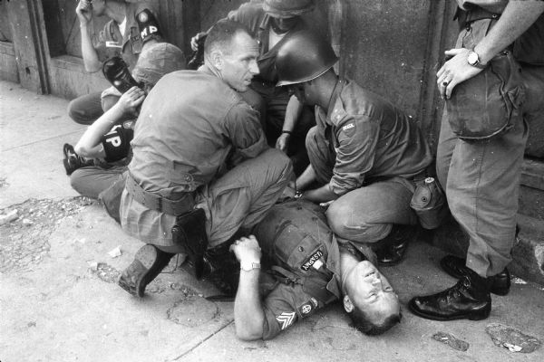 U.S. paratrooper Sgt. Glen Meunier of Port Charlotte, Florida, laying on the ground next to a building after being shot through his right thigh by a rebel in Santo Domingo, Dominican Republic. Meunier is receiving first aid from U.S. military police and a Brazilian officer as he waits for medics.