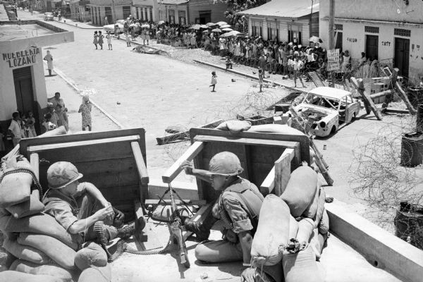 View from rooftop of a blockaded street in Santo Domingo, Dominican Republic, seen from a U.S. Marine position. A large group of civilians, mainly women, some with umbrellas, stand across the street behind coils of barbed wire. A sign posted near the sidewalk nearby reads: "Off Limits, Halt!!, Leaving US Sector."