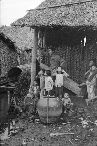 Men and young children in a roofed enclosure outside a dwelling near the village of Binh Hung, South Vietnam. Several of the children are plugging their ears in reaction to the noise of combat. There is boat on a river or canal next to the house on the left.