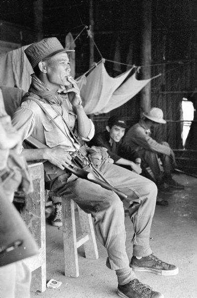 Vietnamese soldier seated indoors on a chair smoking a cigarette. He holds a gun under his right arm. Other men sit behind him.
