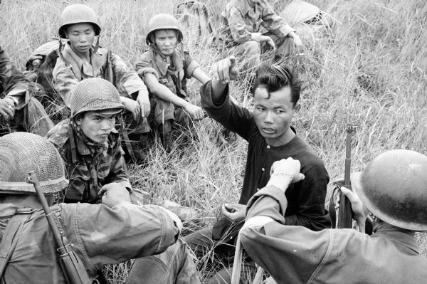 Vietnamese soldier interrogating a prisoner. The prisoner is describing a route of safe passage through the jungle and informing the 1st Vietnamese Marine Battalion where he last saw Viet Cong. Other soldiers observe the interrogation.