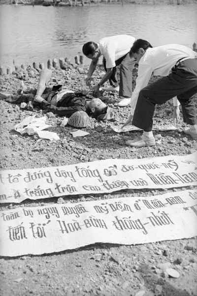 Body of a Viet Cong sympathizer who had been painting anti-government propaganda banners to hang in a South Vietnamese border village. Some of the banners are displayed in the foreground. Two men are standing over him. In the background is a canal.