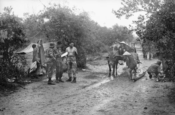 Vietnamese soldiers breaking camp along a muddy road after a night of heavy rain. The shirtless man is battallion commander, Major Giai (?).  Civilian villagers pass on the road with a cart pulled by cattle.