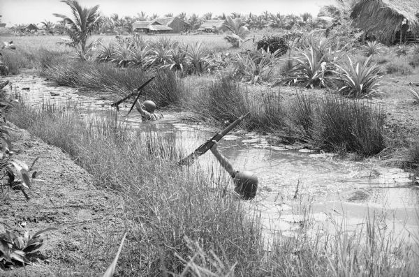 South Vietnamese soldiers from the Sea Swallow Army wading along a canal on patrol near the village of Binh Hung. Grass buildings are in the background near trees.