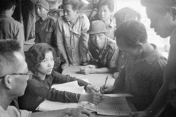 South Vietnamese soldiers gathered around a table where a woman, the "paymistress," sits with payroll sheets. The men are collecting their pay in the village of Binh Hung.