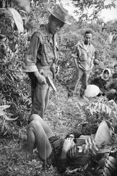 South Vietnamese soldier uses a handgun to threaten a Viet Cong prisoner who is laying on the ground among foliage. Other men watch in the background.