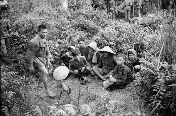 Group of Viet Cong held prisoner by South Vietnamese soldiers. The group is seated on the ground among foliage with mock rifles in the foreground. Soldiers are standing in the far background.