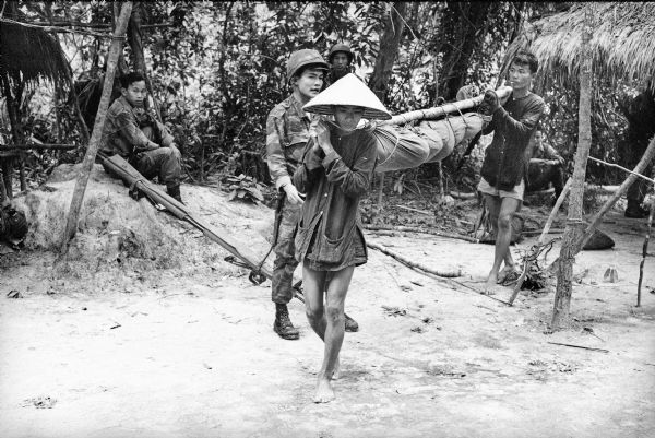 Two Viet Cong prisoners carry the body of a recently killed South Vietnamese paratrooper, wrapped in canvas and tied to a pole. The prisoners are supervised by a South Vietnamese soldier. Other men watch in the background.