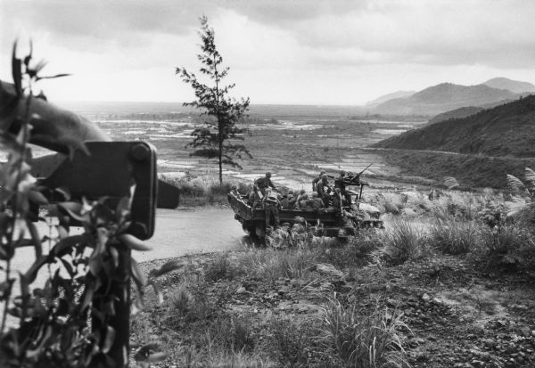 View from roadside of a number of soldiers riding in the back of a military transport truck along a road on a high hillside with a view of the valley below.