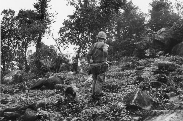 Three soldiers navigate down a rocky hillside covered with low-lying vegetation.