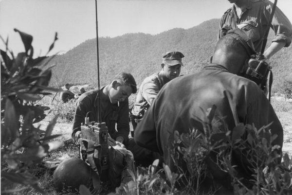 Six soldiers along a roadside in Vietnam. One soldier operates a radio while another writes. Other men sit on the other side of the road, and in the far background are hills.