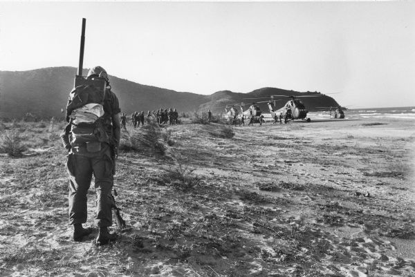 A soldier with a radio on his back waits in the foreground while a group of men stand waiting while other men board helicopters on a beach in Vietnam.