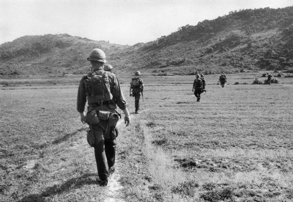 Rear view of group of soldiers walking in an open field on a "search and clear" mission near Da Nang. Hills are in the background.