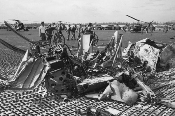 Wreckage of a downed helicopter at an airbase in Vietnam. Several soldiers, other helicopters and tents are in the background.