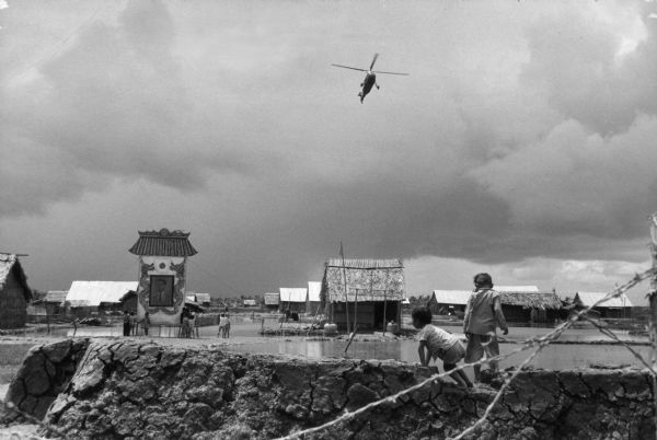 View looking over barbed wire towards a helicopter flying over the village of Binh Hung, a base for the fighting operations for the South Vietnamese. Two children are climbing a mud wall in the foreground near water. More children are standing near a banner on the left.