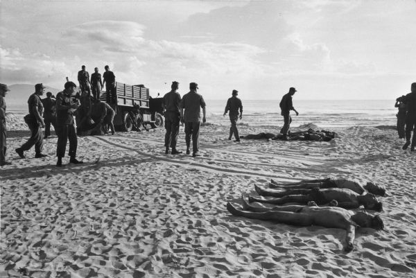 Bodies of several dead Viet Cong soldiers lying on a beach. Several U.S. soldiers observe as bodies are placed in the sand from a truck.