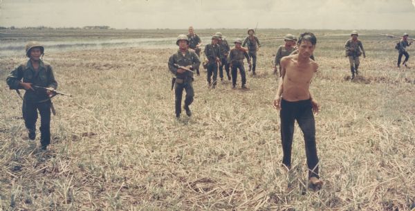 A man caught with Communist propaganda is taken, with his arms bound, to a waiting helicopter. The soldiers who captured him walk behind with weapons raised. A village is in the far background.