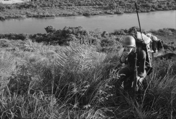View looking down at a U.S. soldier, holding a rifle and carrying a radio unit on his back, sits on a large rock on a ridge overlooking a river.
