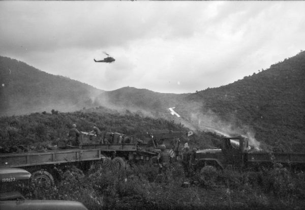A helicopter flies in the sky, with tree-covered hills in the background. In the foreground are several U.S. Marine transport vehicles. Soldiers are working with supplies on the trucks.
