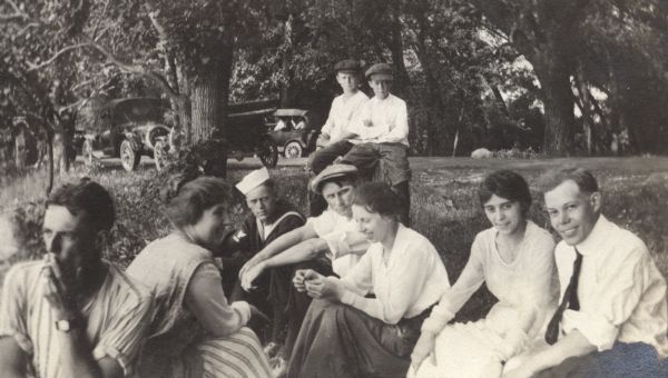 Group of men and women seated in the grass at the side of a road. One man wears a sailor's uniform. Automobiles are parked along the road in the background under large trees.