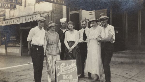 Three couples posed on a sidewalk in front of a Luick Ice Cream sign. An ice cream parlor is in the background. One man is wearing a sailor's uniform.