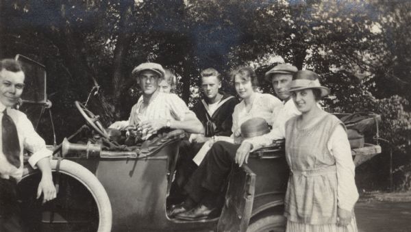 Three couples posed in and in front of an automobile, and another man standing next to the car on the left. Trees are in the background.