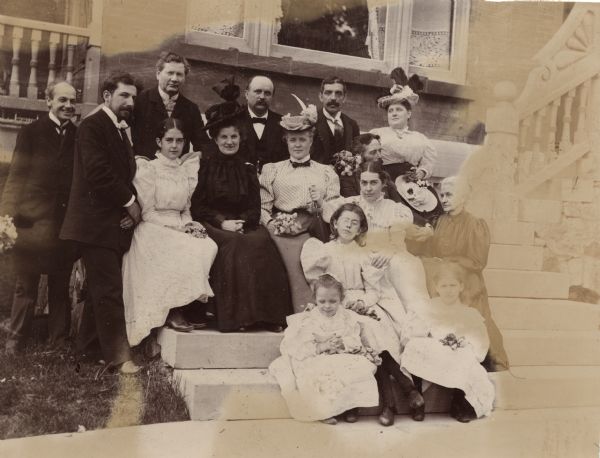 Informal group portrait of fifteen people, men women and children, posed on stone steps at Oakwood Sanitarium. Some of the people are holding bouquets.