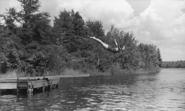 View across water of girl in mid-dive off of diving board. Original caption notes: "Swimming in the clear, cool, PURE waters of our Lake Raymond is a thrill and a pleasure never to be forgotten. Diving, life-saving and water safety, and the harder strokes appeal to good swimmers. Beginners progress beyond the fondest dreams of their parents and of themselves. Long hours of training and experience stand behind those who staff our waterfront. Parents need never worry about their daughter's safety in our care."