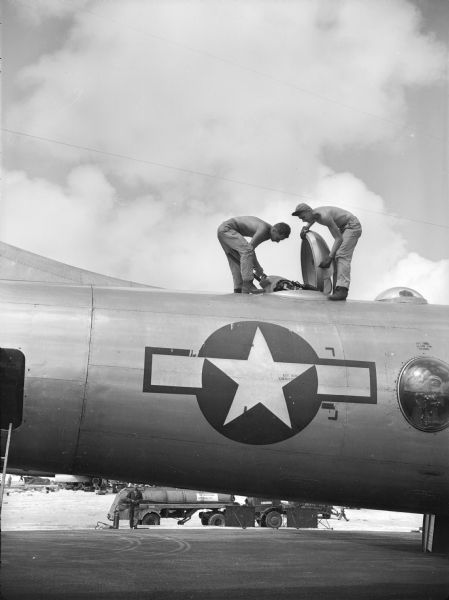 Two men working on a B-29 bomber, just returned from its first fire raid on Tokyo. The men are on top of the plane, opening a hatch. They are both shirtless and one of them is wearing a brimmed cap. The plane has a large star on it with a circle around the star. A tanker truck is in the background.